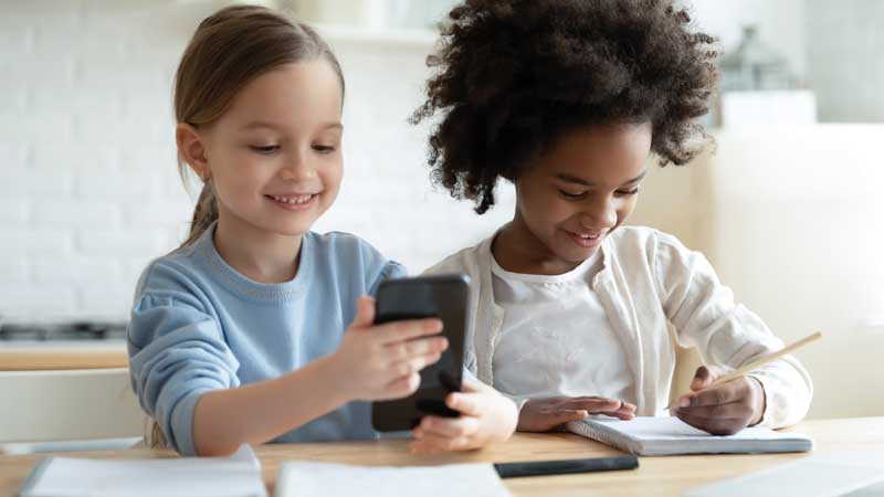 Duas meninas sentadas a uma secretária, a escreverem num caderno e a navegarem num teleóvel